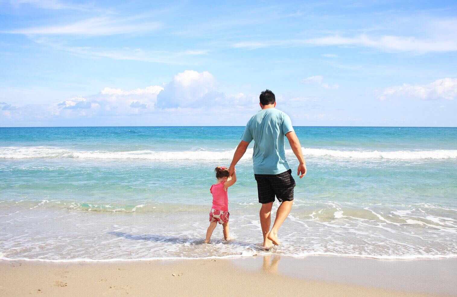 A dad walking hand in hand with his child on a beach.
