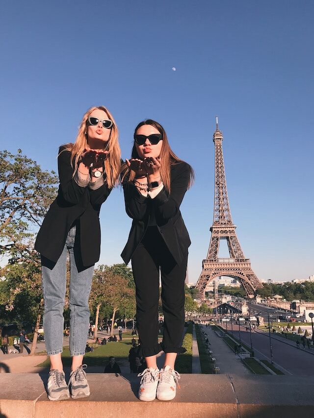 Two women blowing a kiss to the camera. The Eiffel Tower is in the background.