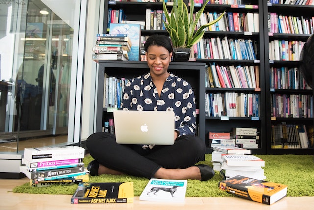 A woman sitting on the floor with her legs crossed. She is surrounded by books and using her laptop.