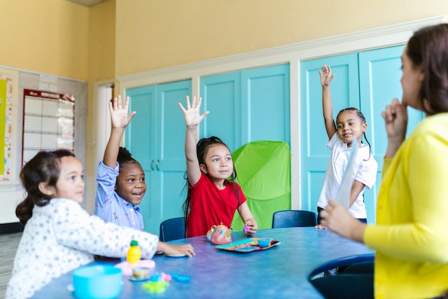 Four children sitting at a long table and raising their hands. The teacher is deciding who to call on.