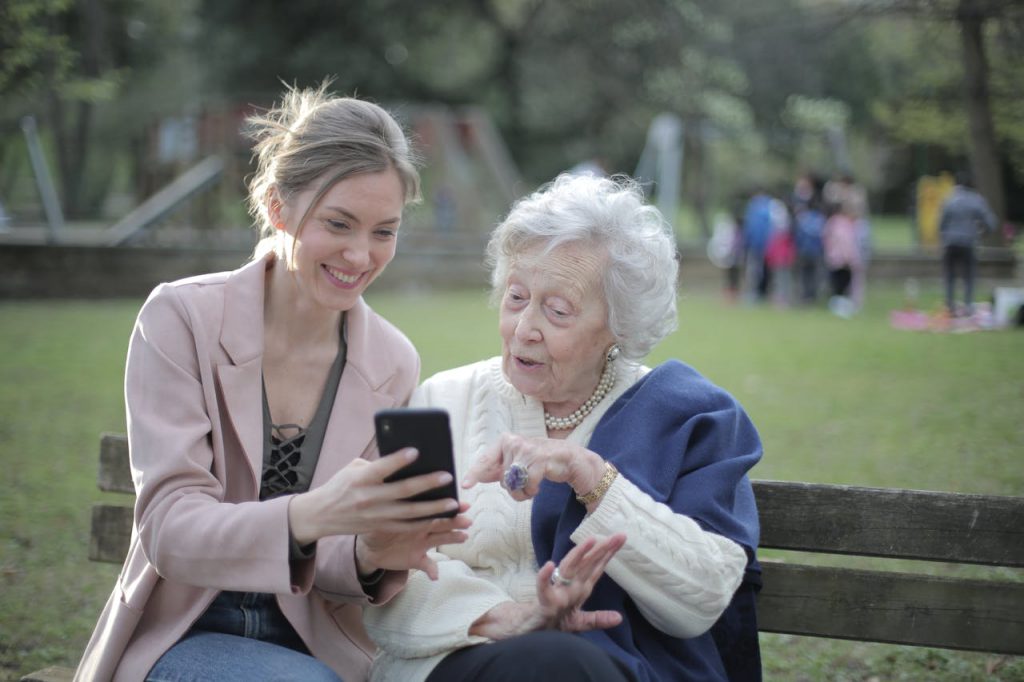 An elderly woman and her adult daughter using a smartphone on a park bench.