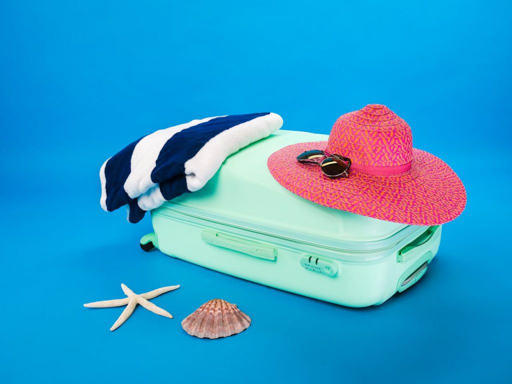 A sun hat and a towel placed on top of a suitcase. A starfish and a seashell are placed next to the suitcase.