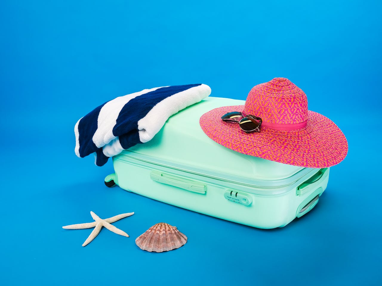 A sun hat and a towel placed on top of a suitcase. A starfish and a seashell are placed next to the suitcase.