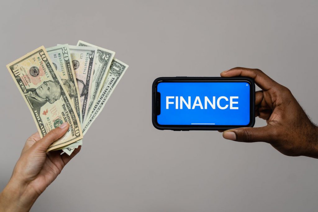 A person holds money in one hand and a smartphone in the other. The smartphone is displaying the word "finance" in large font.