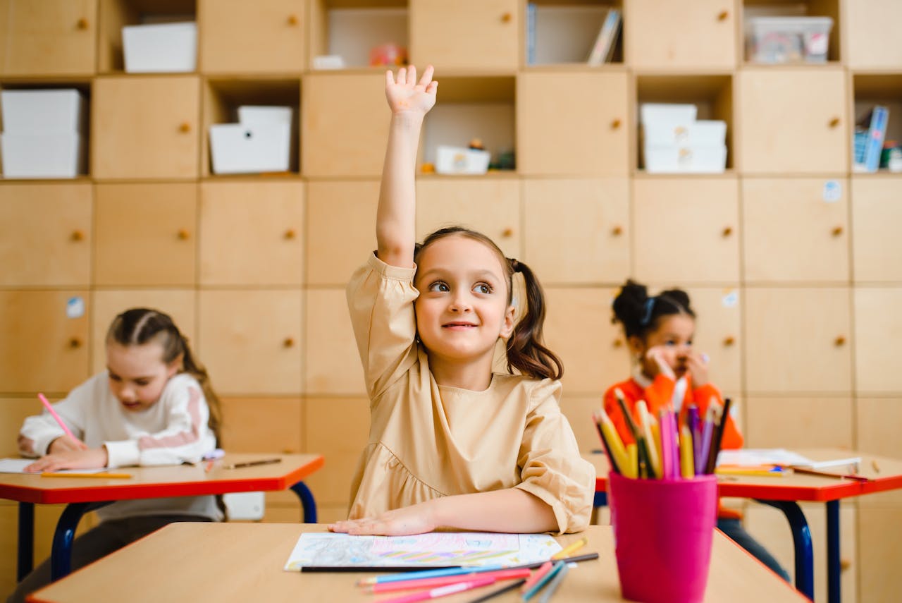 A girl raises her hand while sitting in a classroom with 2 other children.