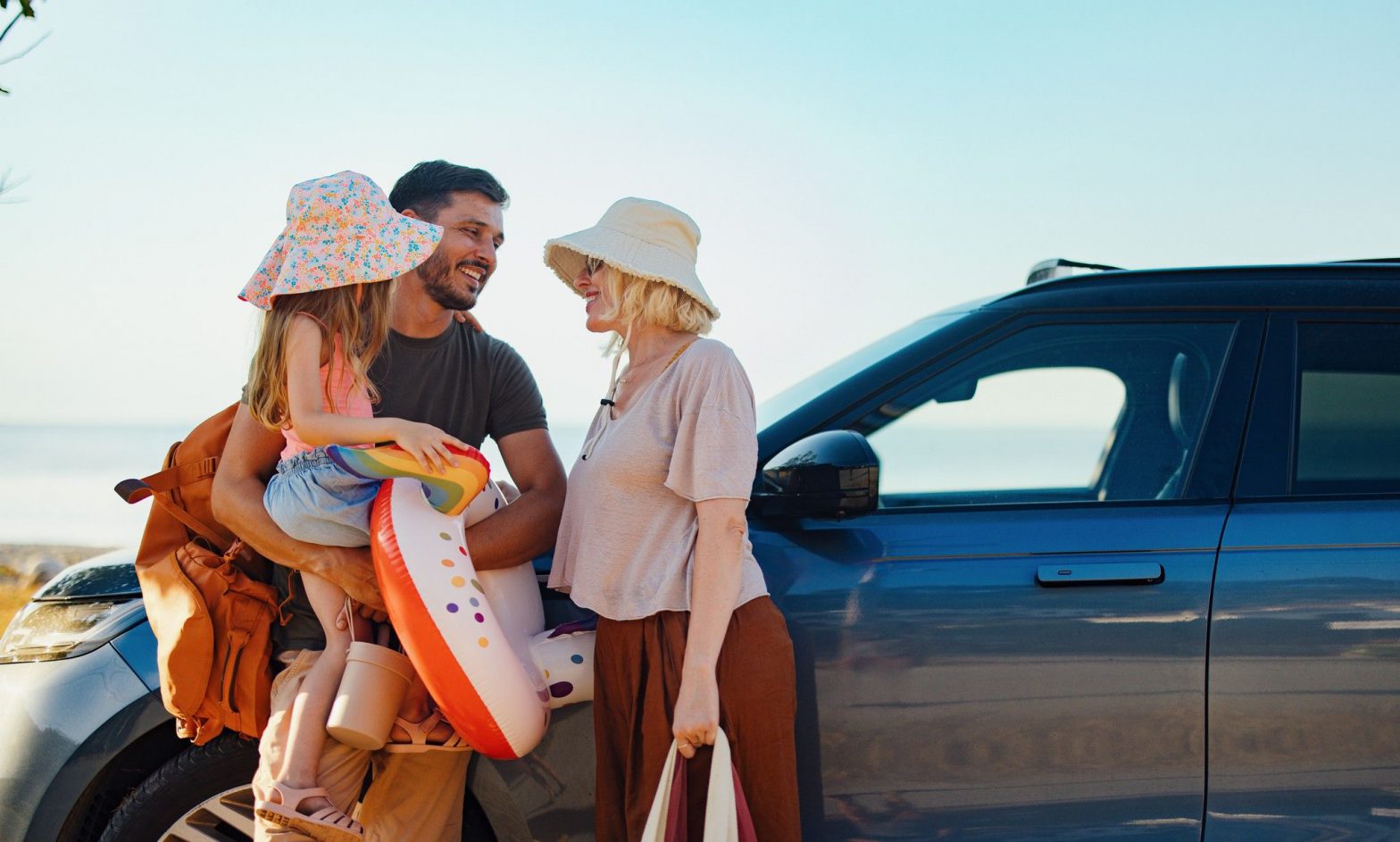 A family gathers by a car. They are by a beach.